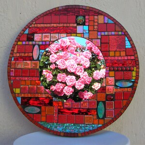 May include: A round mosaic mirror with a red, orange, and blue geometric pattern. The center of the mirror features a reflection of a pink rose bush.