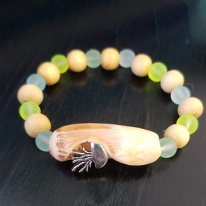 May include: A bracelet made with light brown wooden beads and light blue and green glass beads. A seashell charm with a silver octopus design hangs from the bracelet.