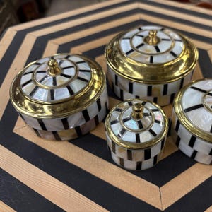 May include: Set of four round decorative boxes with black and white geometric patterns and gold trim. The boxes are made of mother of pearl and have a shiny finish.