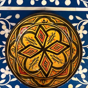 May include: A decorative ceramic plate with a vibrant geometric design. The plate features a yellow center with a black and orange starburst pattern. The rim is black, and the plate is set against a blue background with white floral accents.