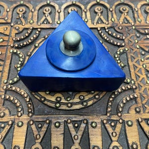 May include: A blue triangular box with a silver knob on the lid. The box is sitting on a wooden surface with intricate carvings.