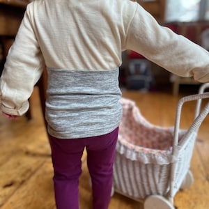 Puede incluir: Un niño pequeño con una camisa de manga larga a rayas crema y gris y pantalones morados está empujando un cochecito de muñecas de mimbre blanco con un forro floral.