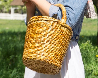 May include: A woman in a denim jacket and white skirt holds a woven wicker basket with a handle. She is holding a bouquet of yellow dandelions in her other hand.