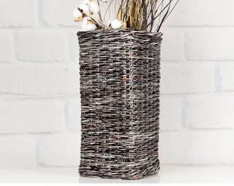 May include: A rectangular woven vase in grey and brown tones, holding dried grasses, golden elements, and cotton stems. The vase is placed on a white surface against a white brick wall, offering a neutral, natural appearance.