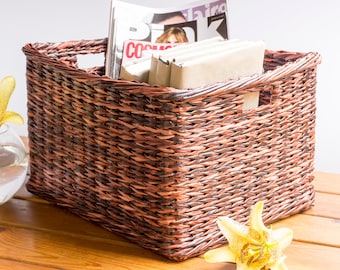May include: A woven wicker basket with a brown and copper finish, filled with magazines and books. The basket is sitting on a wooden table with two yellow lilies.