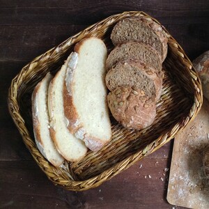 Puede incluir: Una cesta de mimbre llena de rebanadas de pan blanco y una hogaza de pan de masa madre con semillas de calabaza. Una segunda hogaza de pan de masa madre con semillas de calabaza está sobre una tabla de cortar de madera.