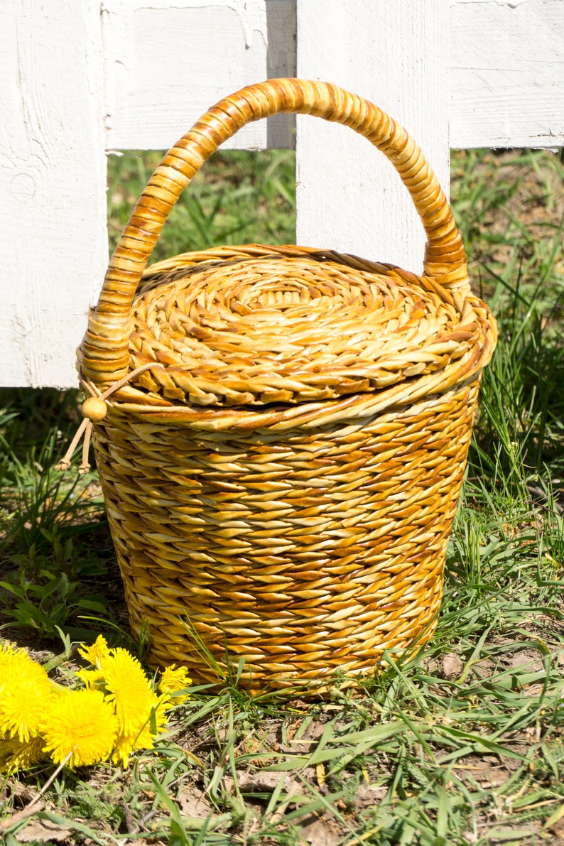 May include: A woven wicker basket with a lid and a handle. The basket is brown and has a natural finish. The basket is sitting on a bed of green grass.