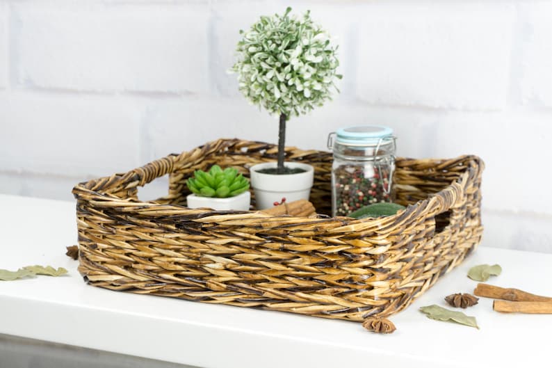 May include: A woven wicker tray with a brown and tan finish. The tray is filled with decorative items, including a small potted succulent, a potted topiary tree, and a glass jar filled with colorful peppercorns.