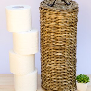 May include: A woven wicker toilet paper holder with a leather handle. The holder is brown and has a natural finish. There are four rolls of white toilet paper stacked next to the holder. A small potted succulent plant is on the right side of the image.