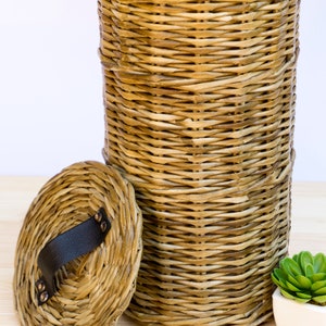 May include: A woven wicker basket with a lid. The lid has a black leather handle with metal rivets. The basket is light brown and has a natural, rustic look.