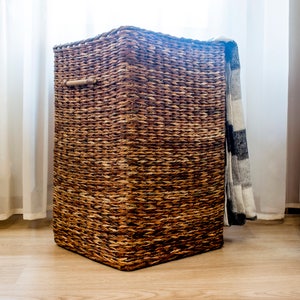 May include: A woven brown wicker storage basket with a wooden handle. The basket is sitting on a wooden floor and has a blanket draped over the top.