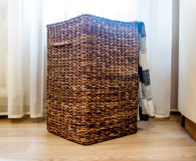 May include: A woven brown wicker storage basket with a wooden handle. The basket is sitting on a wooden floor and has a blanket draped over the top.
