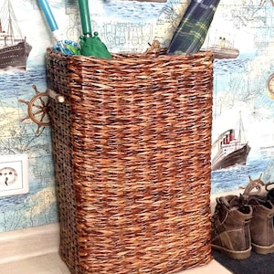 May include: A brown wicker umbrella stand with a green umbrella and two blue umbrellas inside. The stand is sitting on a wooden floor in front of a wall with a nautical themed wallpaper.