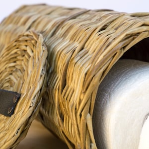 May include: A woven wicker basket with a leather strap handle and a roll of white paper towels inside.