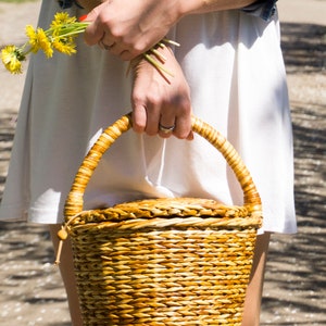 May include: A woman in a white dress and denim jacket holds a woven wicker basket with a handle. The basket is brown and has a natural woven texture.