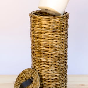 May include: A woven wicker toilet paper holder with a lid and a black leather handle. The holder is filled with a roll of white toilet paper.