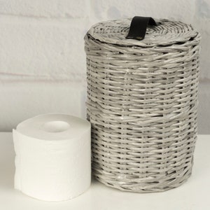 May include: A gray wicker toilet paper holder with a black leather handle. The holder is sitting on a white surface next to a roll of white toilet paper.