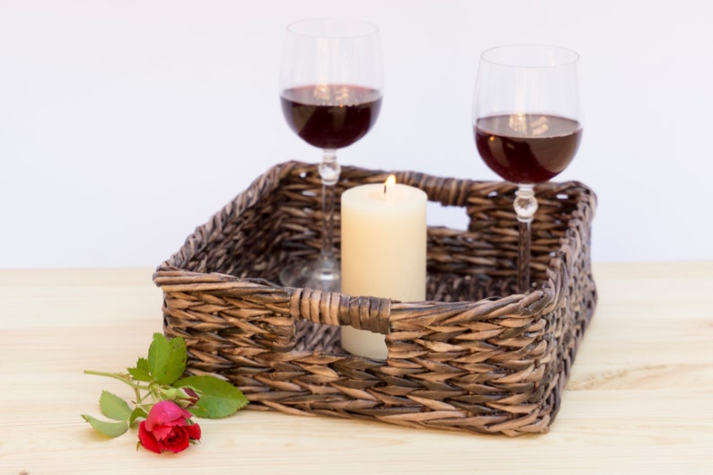 May include: A wicker tray with two glasses of red wine and a lit candle. A single red rose with green leaves is on the table next to the tray.