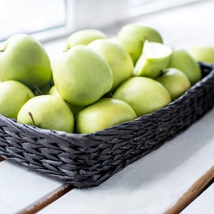 May include: A black woven basket filled with green apples. The apples are arranged in a single layer, with some overlapping. The basket is sitting on a white wooden surface.