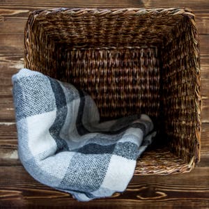 May include: A woven wicker basket with a gray and white plaid blanket inside. The basket is sitting on a wooden surface.