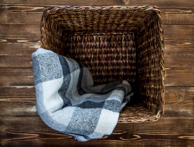 May include: A woven wicker basket with a gray and white plaid blanket inside. The basket is sitting on a wooden surface.
