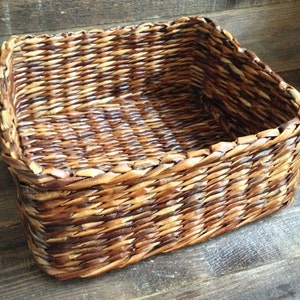 May include: A square woven basket made of natural brown and light brown materials. The basket is empty and has a smooth, woven texture.