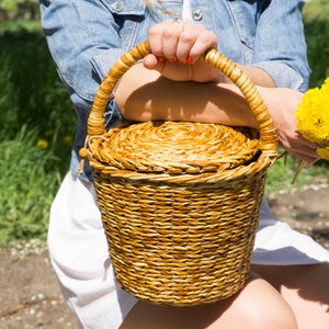 May include: A woman is holding a woven wicker basket with a lid. The basket is brown and has a natural finish. The woman is wearing a denim jacket and a white dress. She is holding a bouquet of yellow dandelions in her other hand.