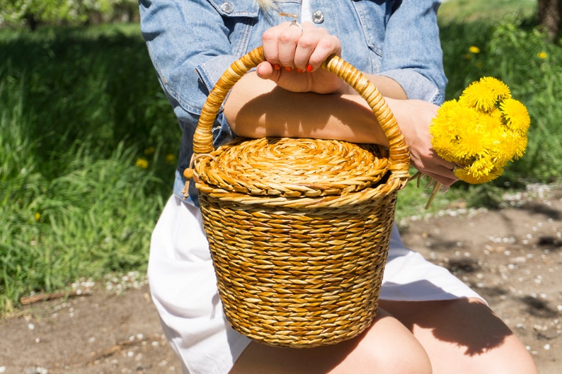 May include: A woman is holding a woven wicker basket with a lid. The basket is brown and has a natural finish. The woman is wearing a denim jacket and a white dress. She is holding a bouquet of yellow dandelions in her other hand.
