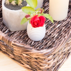 May include: A brown wicker basket with a white ceramic vase with a red rose inside, a white ceramic pot with a small green plant, and a white candle.