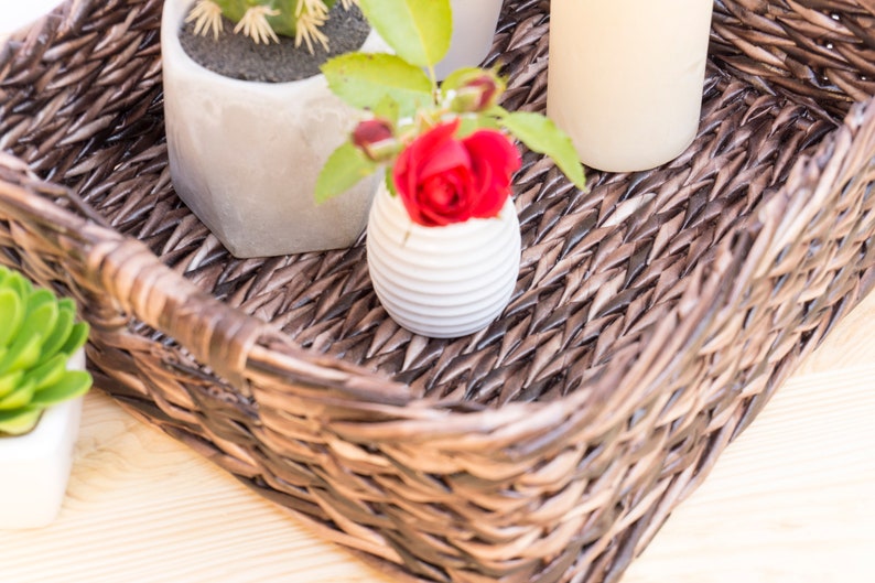 May include: A brown wicker basket with a white ceramic vase with a red rose inside, a white ceramic pot with a small green plant, and a white candle.