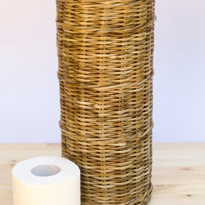 May include: A woven wicker toilet paper holder with a black leather handle and a roll of white toilet paper in front of it.