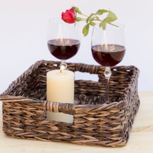 May include: A wicker tray with two glasses of red wine, a lit candle, and a single red rose with green leaves.
