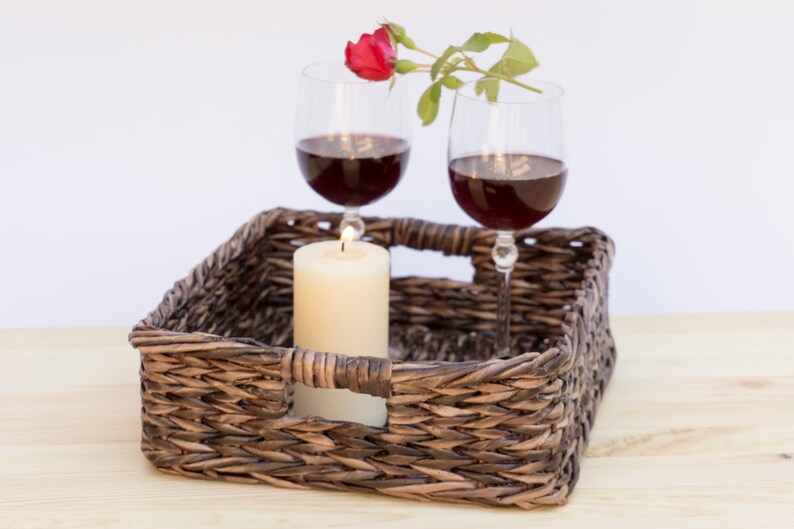 May include: A wicker tray with two glasses of red wine, a lit candle, and a single red rose with green leaves.