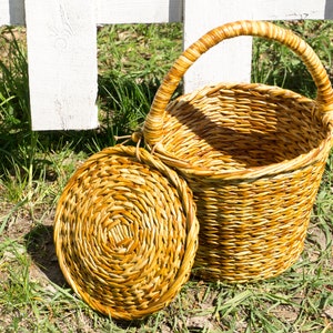 May include: A woven wicker basket with a handle and a matching lid. The basket is light brown and has a natural, rustic look.