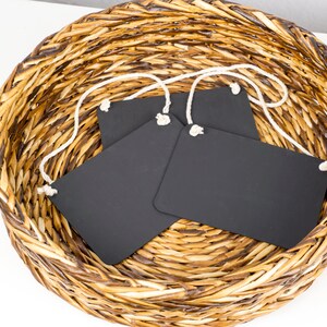 May include: Three black chalkboard tags with white string attached, sitting in a woven wicker basket.