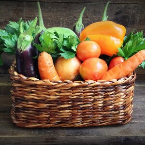 May include: A wicker basket filled with fresh produce, including carrots, tomatoes, onions, a bell pepper, and eggplant. The basket is sitting on a wooden surface.