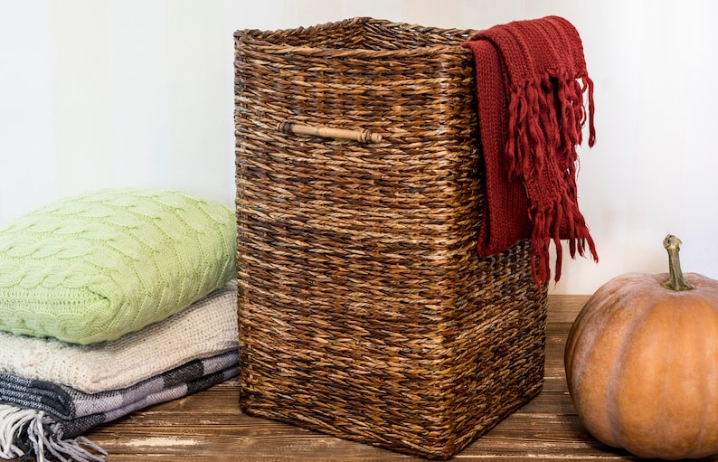 May include: A woven wicker storage basket with a wooden handle. The basket is filled with a red knitted throw blanket and is sitting on a wooden surface next to a pumpkin.