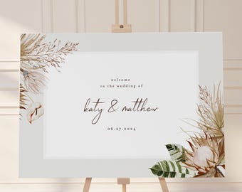 May include: Welcome sign with a light gray background and brown text. The sign says "Welcome to the wedding of Katy & Matthew 06.27.2024". The sign is decorated with dried floral arrangements in shades of brown, beige, and green.