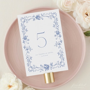 Puede incluir: Una tarjeta de número de mesa blanca con un borde floral azul y el número 5 en azul. El texto "La Tourelle Hotel 9.18.2026" está impreso en azul en la parte inferior de la tarjeta.