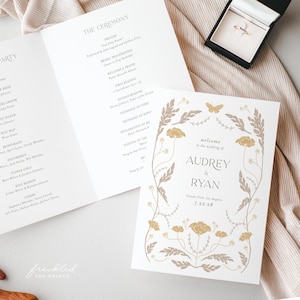 May include: Wedding stationery set featuring a program and invitation. The invitation has a floral design with the names "Audrey & Ryan" and the wedding date. A ring is displayed in a black box.