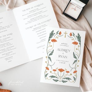 May include: Wedding stationery set featuring a program and invitation. The invitation has a floral design with orange flowers and the names Audrey & Ryan. The program details the ceremony. A ring box is visible.