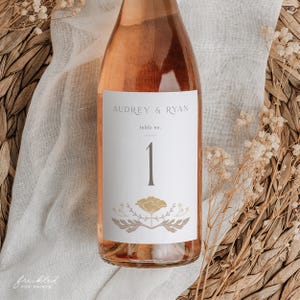 May include: A pink wine bottle with a white label that says "Audrey & Ryan" and "table no. 1". The label has a floral design with a poppy.