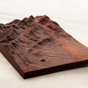 May include: A dark brown wooden relief map featuring a carved mountain range topography. The surface displays engraved geographic coordinates reading 37.920068N, 105.412257W and a small star symbol etched into the lower section of the wooden block.