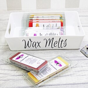 May include: A white plastic container with a black label that says "Wax Melts". The container is filled with small, clear plastic containers of wax melts in various colors. Two wax melts are outside the container, one is red and the other is yellow.