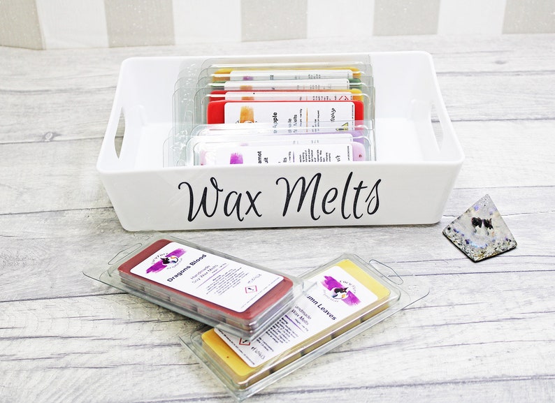 May include: A white plastic container with a black label that says "Wax Melts". The container is filled with small, clear plastic containers of wax melts in various colors. Two wax melts are outside the container, one is red and the other is yellow.
