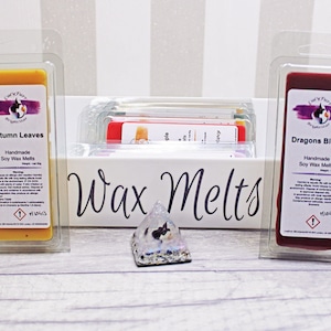 May include: Two soy wax melts in clear plastic packaging. The wax melt on the left is yellow and labeled "Autumn Leaves". The wax melt on the right is red and labeled "Dragons Blood". Both wax melts are handmade and have a warning label.