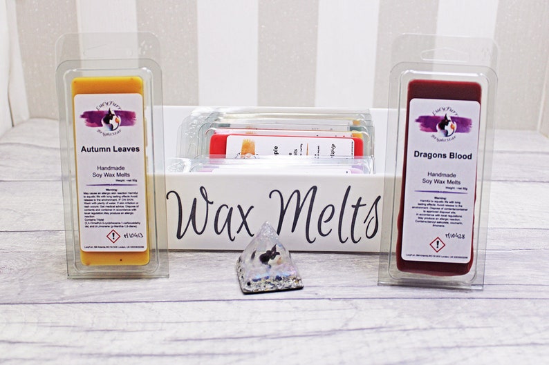 May include: Two soy wax melts in clear plastic packaging. The wax melt on the left is yellow and labeled "Autumn Leaves". The wax melt on the right is red and labeled "Dragons Blood". Both wax melts are handmade and have a warning label.