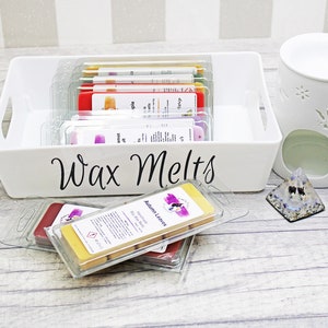 May include: A white ceramic wax warmer with a floral design, a white tray labeled "Wax Melts" filled with various wax melts, and a clear glass pyramid with a blue and white design.