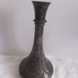 May include: A tall, slender metal vase with a dark, oxidized finish featuring intricate, etched floral patterns throughout the body. The vase has a flared rim, a narrow neck with a decorative ring detail, and a wide, rounded base.