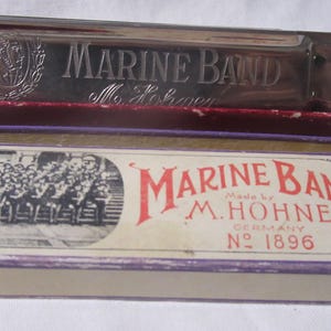 May include: A vintage Marine Band harmonica in its original box. The silver harmonica is engraved with "MARINE BAND" and "M. Schorer". The box features the text "Marine Band Made by M. Hohner Germany No. 1896" and a black and white image of a band.